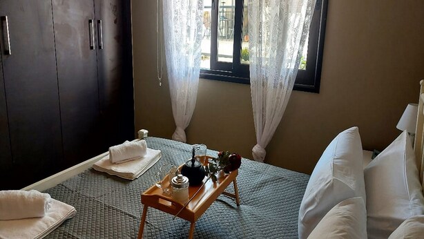 A bedroom with a bed, a bedside table with a lamp, a window with curtains, and a wardrobe. - Paramount Gardens Resorts C202 (Tersefanou)