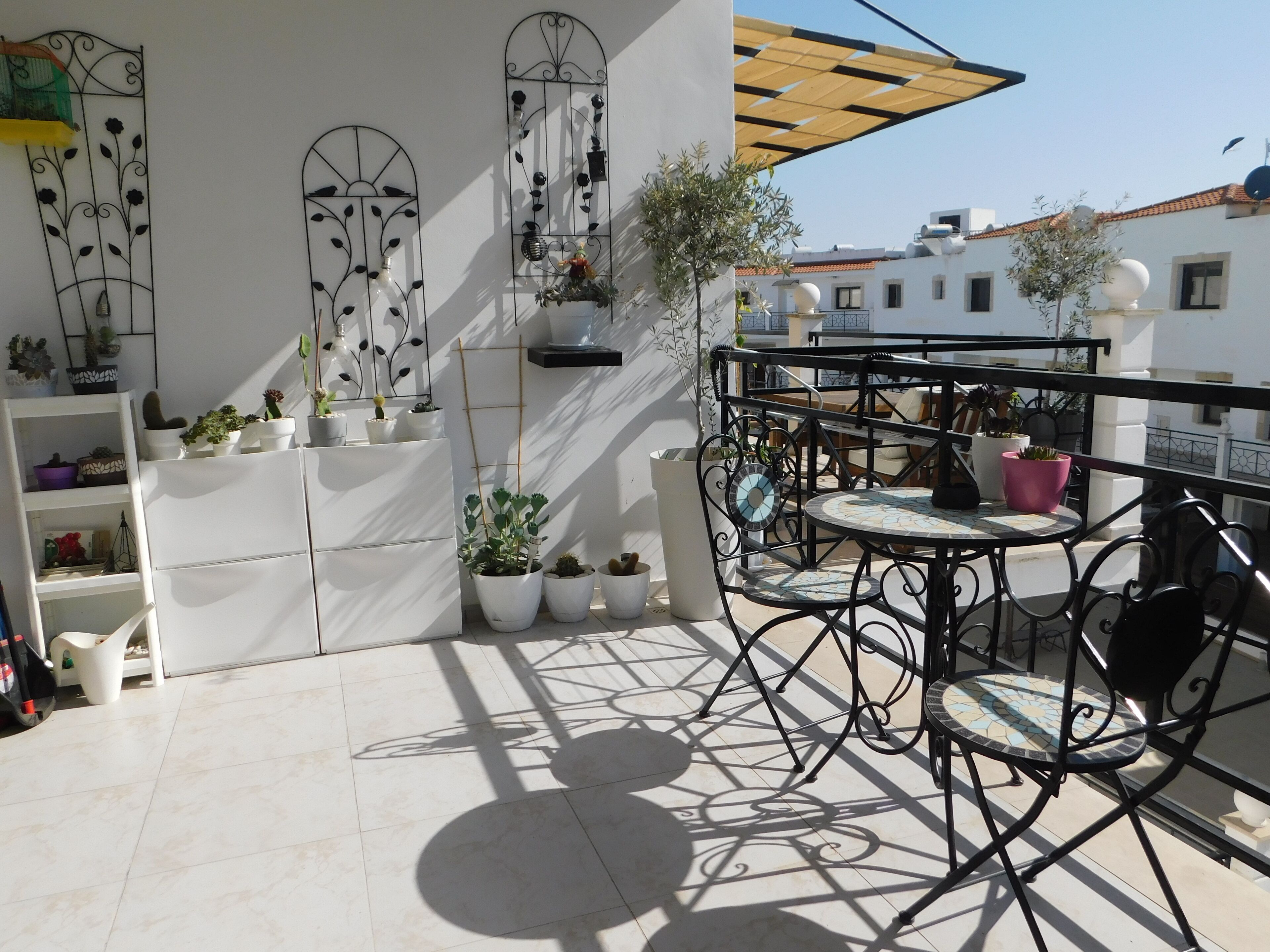 A sunlit outdoor patio with a round metal table and chairs, surrounded by potted plants and decorative metalwork.