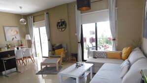 A modern living room with a sofa, coffee table, and chairs. There is a dining area with a table and chairs. A large window provides natural light and a view of the outdoors. - Paramount Gardens Resorts C202 (Tersefanou)