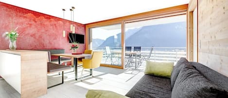 A modern living room with a sofa, a dining area with a table and chairs, a kitchen area with a counter and hanging lights, and a large window offering a view of the mountains.