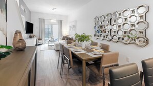Et moderne spiseområde med et træbord dækket til fire personer, en stor sekskantet spejlvæg, og udsigt til opholdsstuen gennem en glasdør.