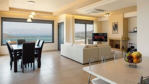 Uma sala de estar moderna com área de jantar, janelas amplas com vista, lareira e uma tela de TV exibindo 'NETFLIX'.