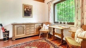 A room with a wooden cabinet, a chair, a table, a window with curtains, and a patterned rug. - Apartment Sassella - Mäser-Apartments (Tschagguns)