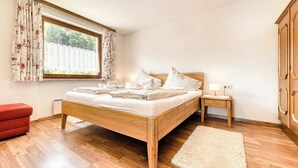 A bedroom with a wooden bed, a nightstand with a lamp, a red chair, and a window with floral curtains. - Apartment Sassella - Mäser-Apartments (Tschagguns)