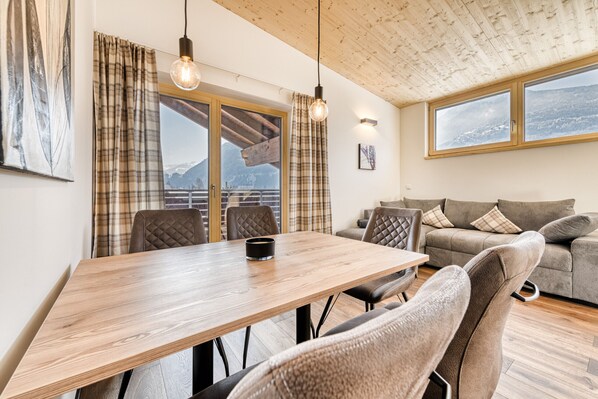 Una sala de estar moderna con una mesa de comedor de madera, dos sillas, un sofá y vistas a las montañas a través de grandes ventanales.