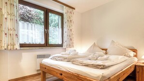 A wooden bed with white linens, a window with floral curtains, and a bedside table with a lamp. - Appartment Golm - Mäser-Apartments (Tschagguns)