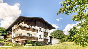 A multi-story chalet with wooden balconies, surrounded by lush greenery and a clear sky.