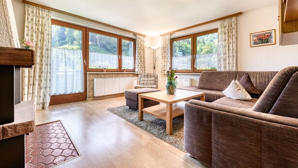A cozy living room with a sofa, a wooden coffee table, and a chair. There are large windows with curtains, a radiator, and a framed picture on the wall.