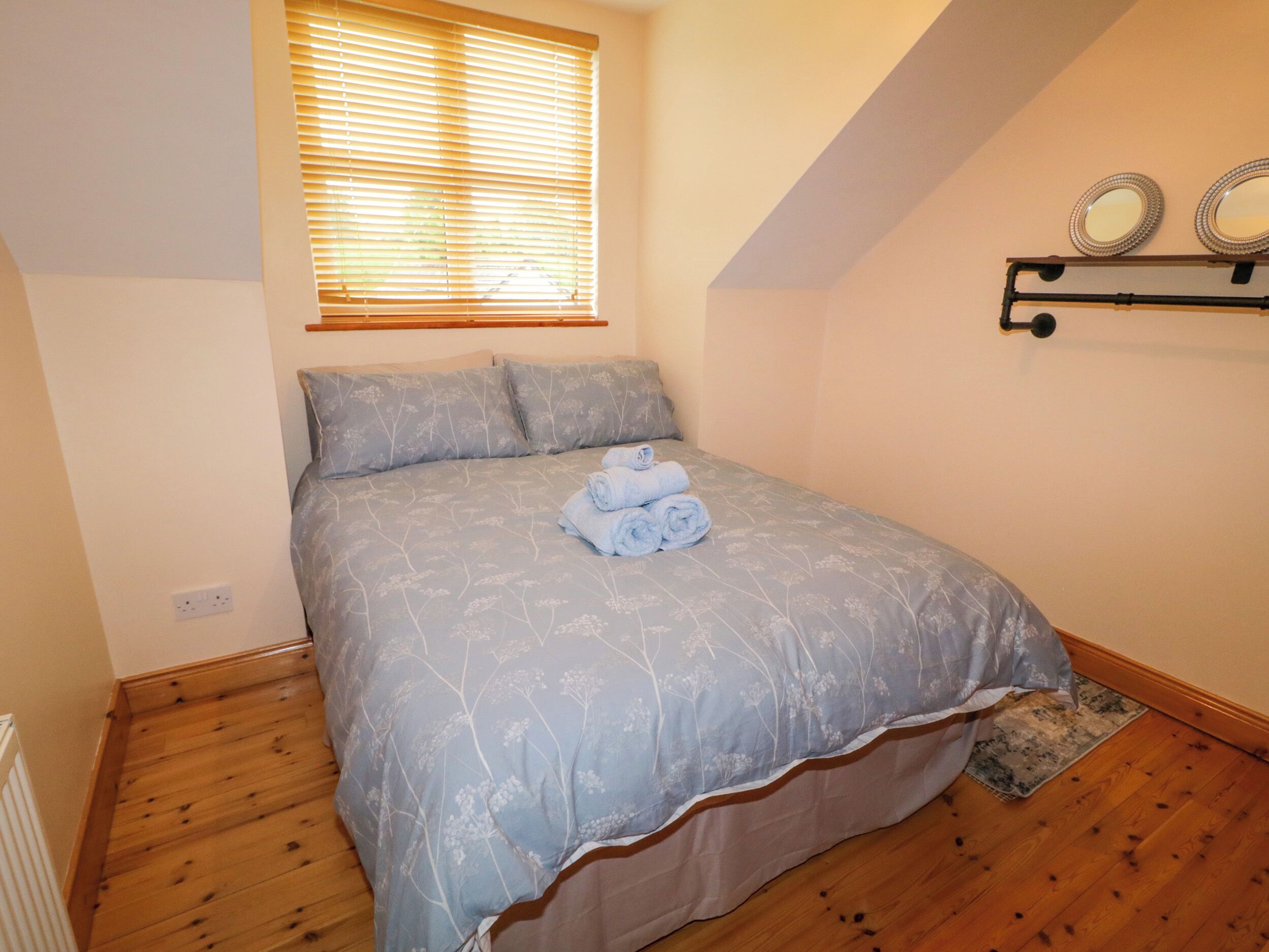 Une chambre à coucher avec un lit, une fenêtre avec des stores, une étagère fixée au mur avec deux miroirs et un petit tapis.