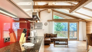 A modern kitchen with a central island, a dining area with a wooden table and chairs, and a large sliding glass door leading to a balcony with a view of trees.