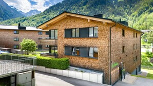 Ein modernes, zweistöckiges Backsteingebäude mit großen Fenstern und einem Balkon, umgeben von üppigem Grün und Bergen.