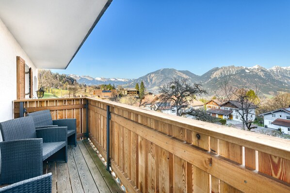 A balcony with wicker furniture, a wooden railing, and a view of mountains and houses.