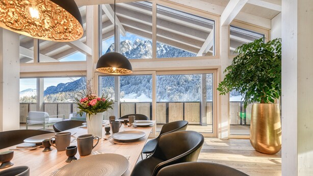 Comedor con una mesa de madera dispuesta para seis comensales, ventanas amplias con vista a la montaña y una planta grande en maceta.