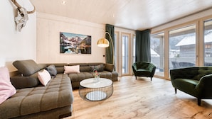 Ein modernes Wohnzimmer mit Holzbalkendecke, einem großen Fenster, einem grünen Sessel, einer Couch und einem runden Couchtisch.