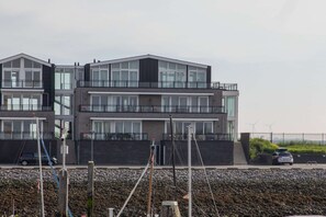 Un moderno edificio de varios pisos con amplios ventanales y balcones, ubicado cerca de una costa rocosa con aerogeneradores al fondo.