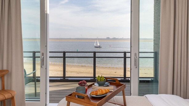 Varanda com mesa posta, exibindo um croissant, uvas e uma bebida, com vista para uma praia e um veleiro ao longe, além de turbinas eólicas.