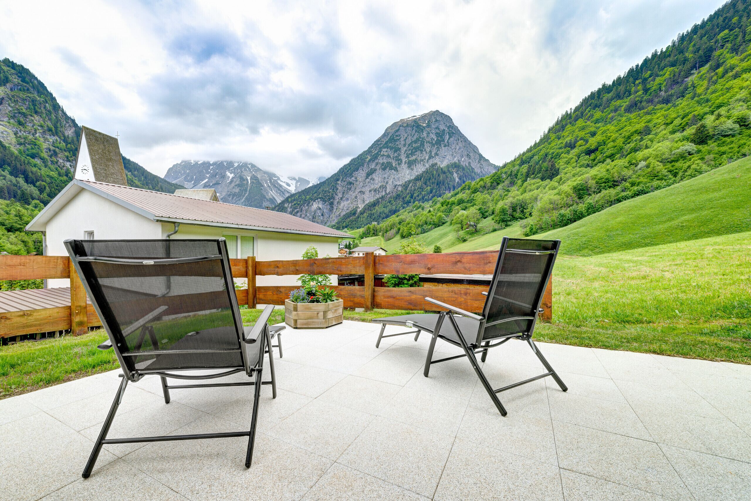 Une terrasse avec deux chaises noires et une table, une clôture en bois et un bâtiment à la façade blanche, orné d’une horloge.