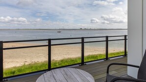 Um deck de madeira com vista para uma praia e um corpo d'água, com guarda-corpo e uma mesa.