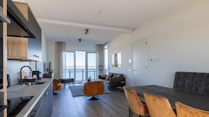 Cozinha moderna com ilha central, sala de jantar com mesa e cadeiras, e sala de estar com sofá e mesinha de centro.