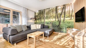 A modern living room with a large sectional sofa, a wooden coffee table, and a view of a forest through a large window. - Holiday apartment Aurora (St. Gallenkirch)