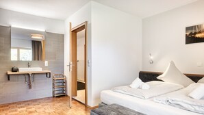 A modern hotel room with a wooden floor, a large bed with white linens, a bedside table, a built-in bench, a large mirror, a vanity with a sink, and a wooden door leading to a closet. - Holiday apartment Aurora (St. Gallenkirch)