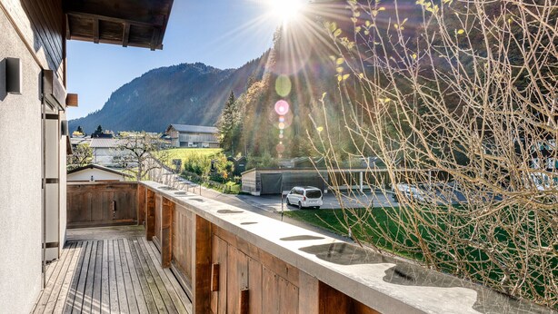 A wooden deck with a stone ledge, overlooking a mountain and a road with a white van. - Holiday apartment Aurora (St. Gallenkirch)