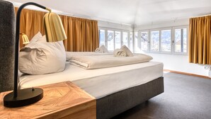 Una camera d'albergo moderna con un grande letto, una scrivania con una lampada e ampie finestre che offrono vista sulle montagne.