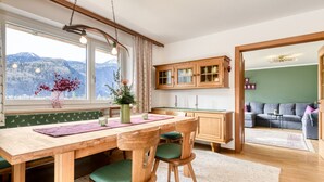 Un coin repas avec une table et des chaises en bois, un vase rempli de fleurs et une vue sur les montagnes, visible à travers de grandes fenêtres.