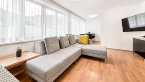 Un salon moderne avec un canapé d’angle gris, une table basse en bois, un coussin jaune, un téléviseur à écran plat et une grande fenêtre ornée de rideaux légers.