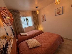 Une chambre avec deux lits, une fenĂȘtre avec des rideaux, un luminaire suspendu et des Ćuvres dâart encadrĂ©es accrochĂ©es au mur.