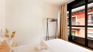 A hotel room with a bed, a window with a view of buildings, a metal rack, and a vase with dried flowers. - Apartamentos Aptio 644C3 (Oviedo)
