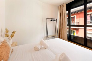 A hotel room with a bed, a window with a view of buildings, a metal rack, and a vase with dried flowers.
