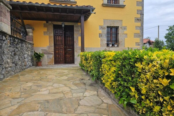 Una entrada empedrada con puerta de madera, un edificio amarillo con detalles de piedra y un seto con flores amarillas.