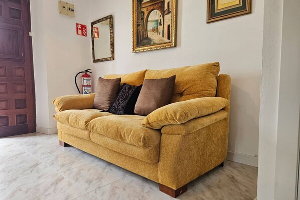 A yellow sofa with multiple cushions, a fire extinguisher, and framed artwork on the wall.