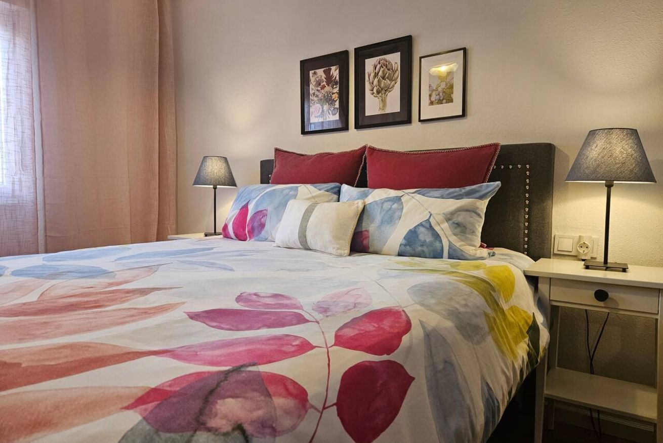 Un lit recouvert d’une couvre-lit à motifs floraux, deux lampes de chevet et trois tableaux encadrés accrochés au mur.