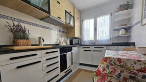 A kitchen with white cabinets, a microwave, a stove, and a sink. There is a window with a curtain, a basket with plants, and a table with a floral tablecloth. - 645A Coqueto apartamento en Oviedo (Oviedo)
