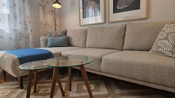 Een woonkamer met een beige bank, een glazen salontafel, een kamerkleed met een patroon, en twee ingelijste kunstwerken aan de muur.
