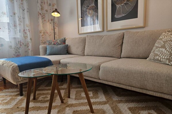 A living room with a beige sofa, a glass coffee table, a patterned rug, and two framed wall art pieces.