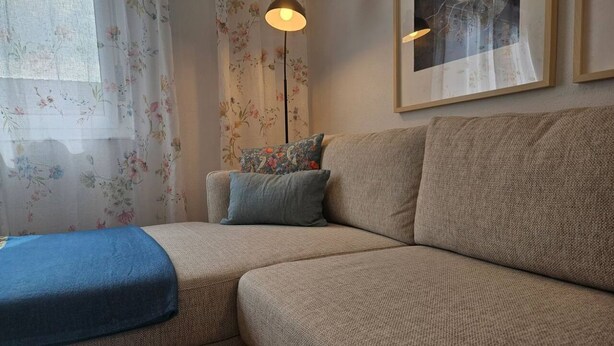 Una sala de estar con un sofá, una manta azul, una lámpara de pie y un cortinaje floral.