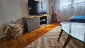 Una sala de estar con una televisión de pantalla plana sobre un mueble de madera, una mesa de vidrio, una cesta de mimbre y un tapete estampado.