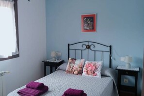 Une chambre à coucher avec un lit, deux tables de chevet, une commode, une fenêtre avec des rideaux et un tableau encadré au mur.