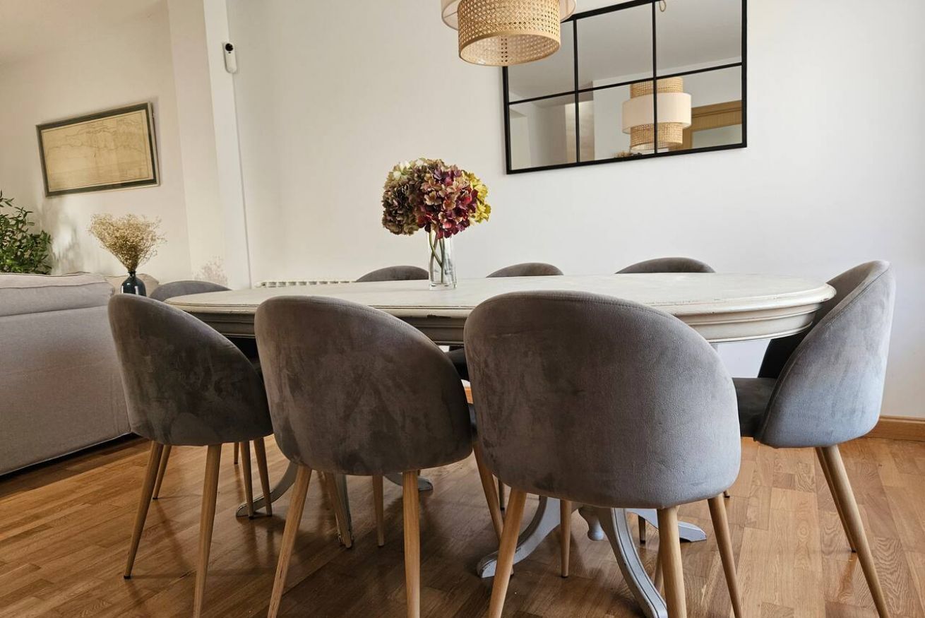 Un comedor con una mesa redonda, sillas grises tapizadas, un florero con flores, una lámpara colgante y un espejo enmarcado en la pared.