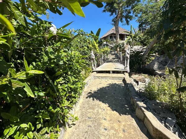 A stone pathway leading to a thatched-roof building surrounded by lush greenery.