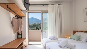 A bedroom with a bed, a TV mounted on a wooden shelf, a bedside table, and a view of the outdoors. - KaYa SeaView Residence (Mylopotamos)