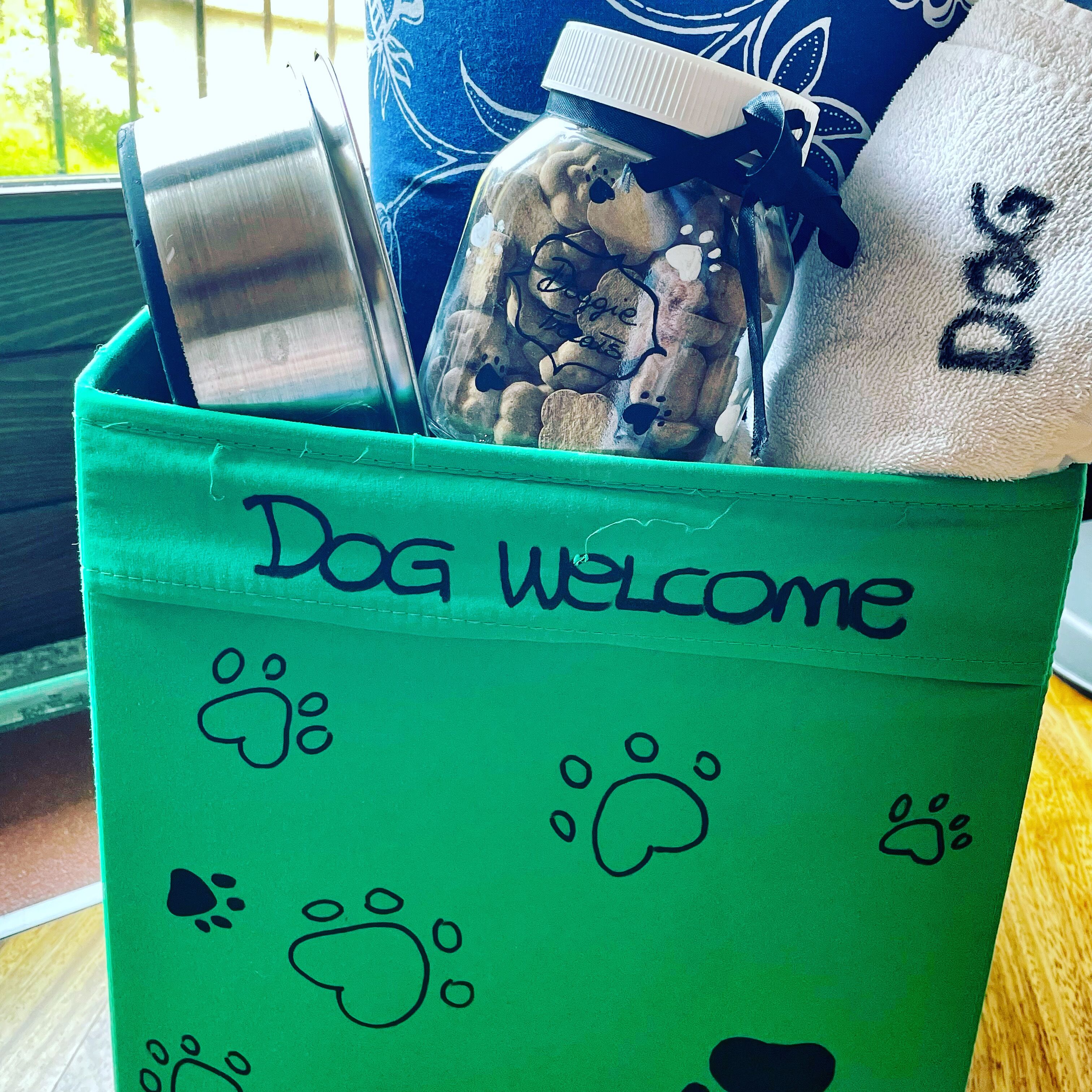 A green storage bin with the text "Dog Welcome" and paw prints, containing a blue pillow, a white towel, and a glass jar with rocks.