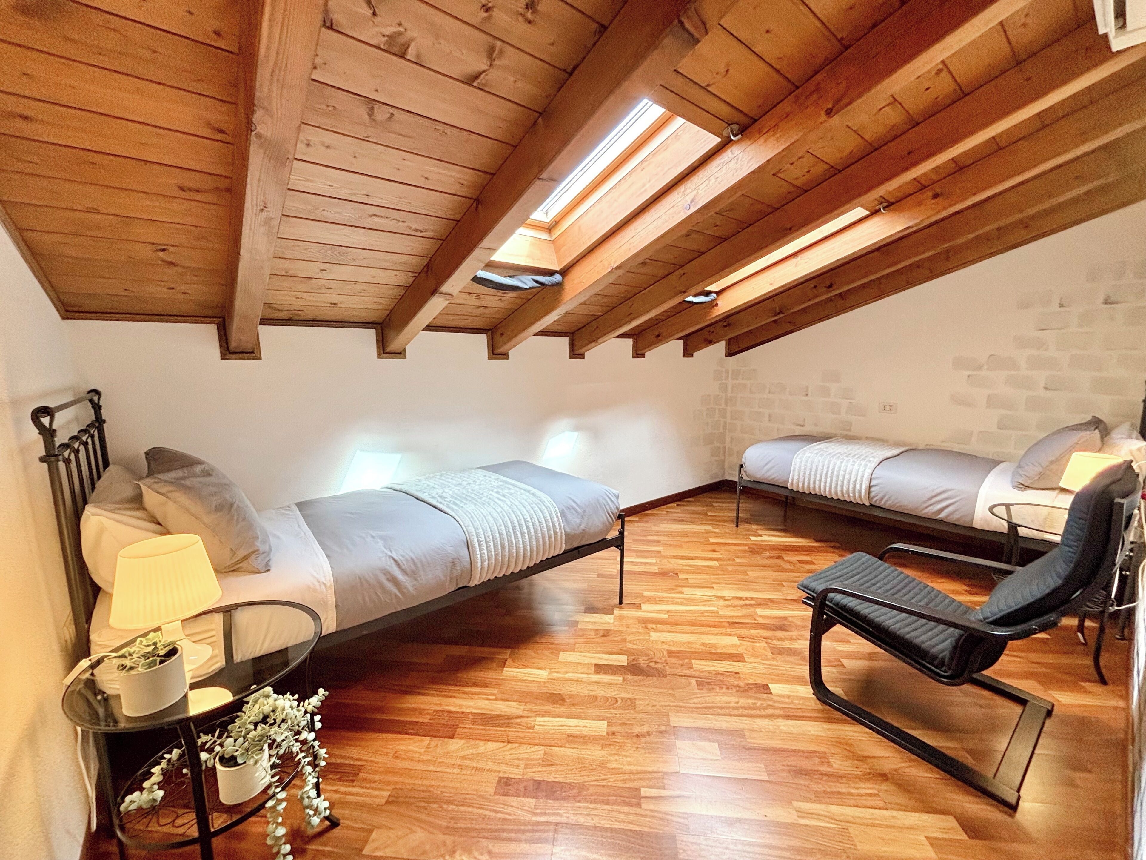A loft-style bedroom with two beds, a black chair, a small table with a plant, and wooden flooring.