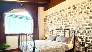 Een slaapkamer met een metalen bedframe, een houten vloer, een wand met baksteenmotief, een raam met gordijnen en een nachtkastje met een lamp.