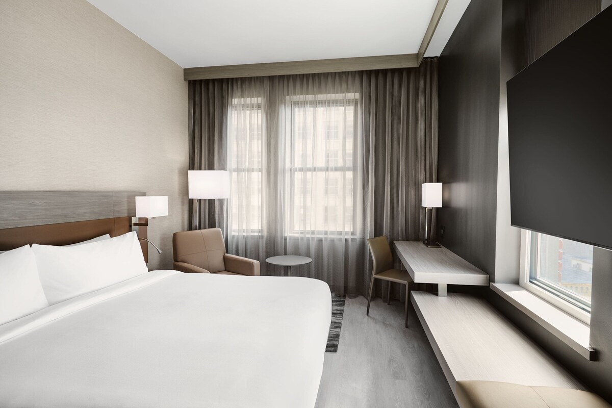 Quarto de hotel com uma cama grande, duas poltronas, uma mesa redonda pequena, uma escrivaninha e uma televisão.