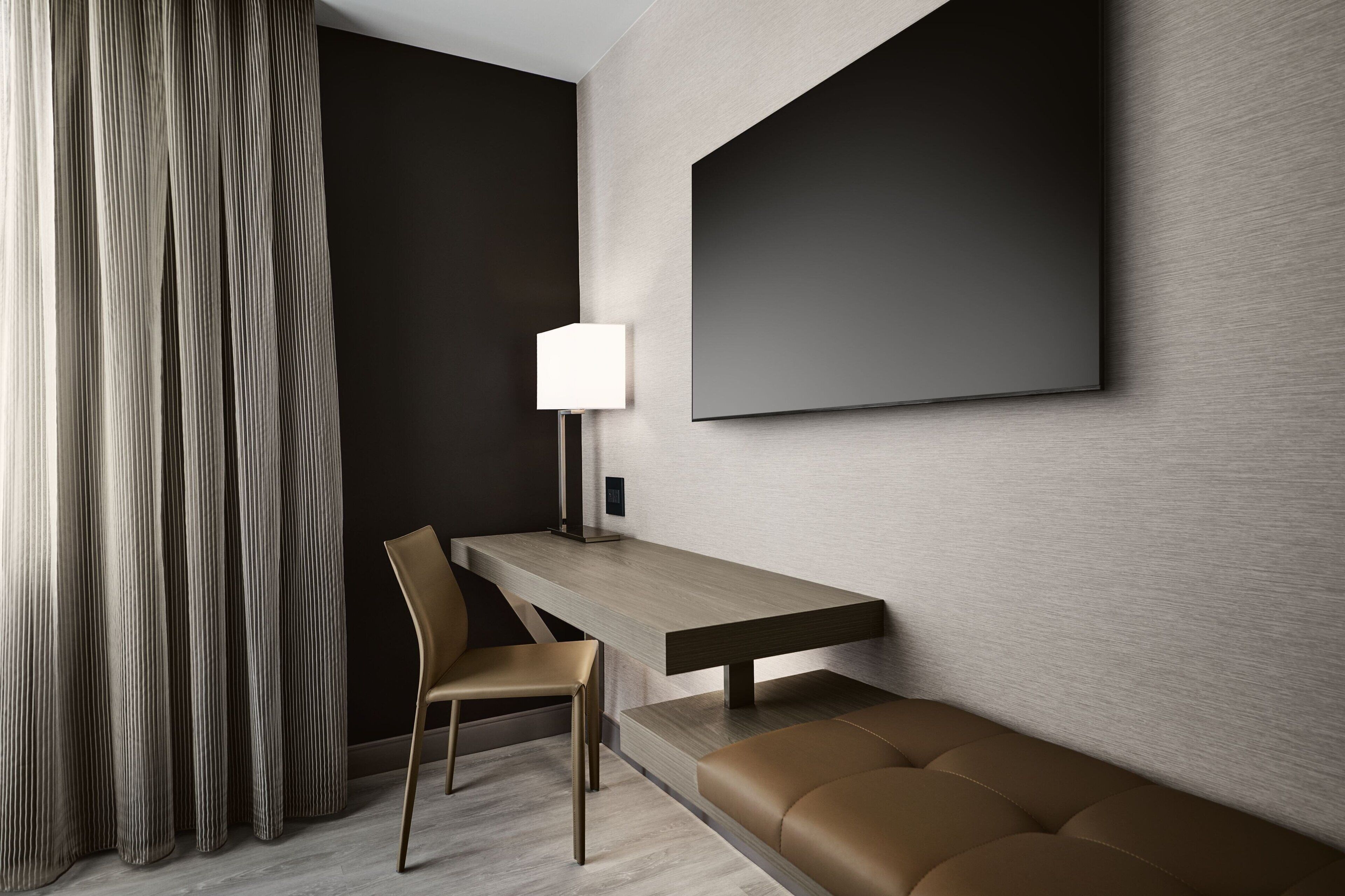 A modern hotel room with a wooden desk, a chair, a bench, a large wall-mounted TV, and a lamp.