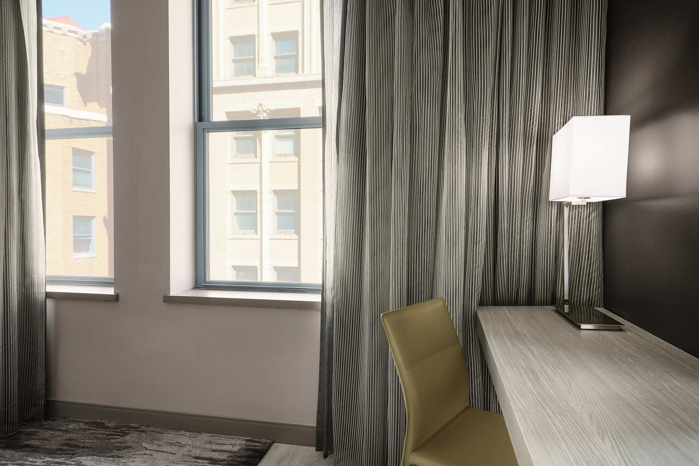 A hotel room with a large window, a desk, a chair, and a lamp.